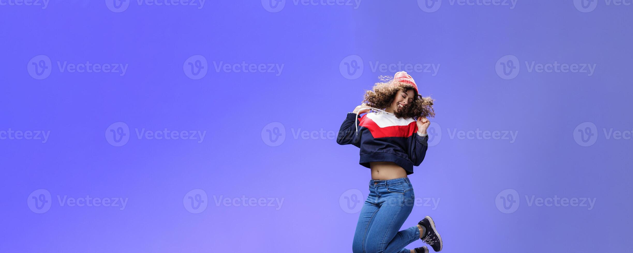 Girl jumping from happiness and delight feeling carefree close eyes and smiling broadly enjoying moment having fun raising hands being dreamy and playful over blue background photo