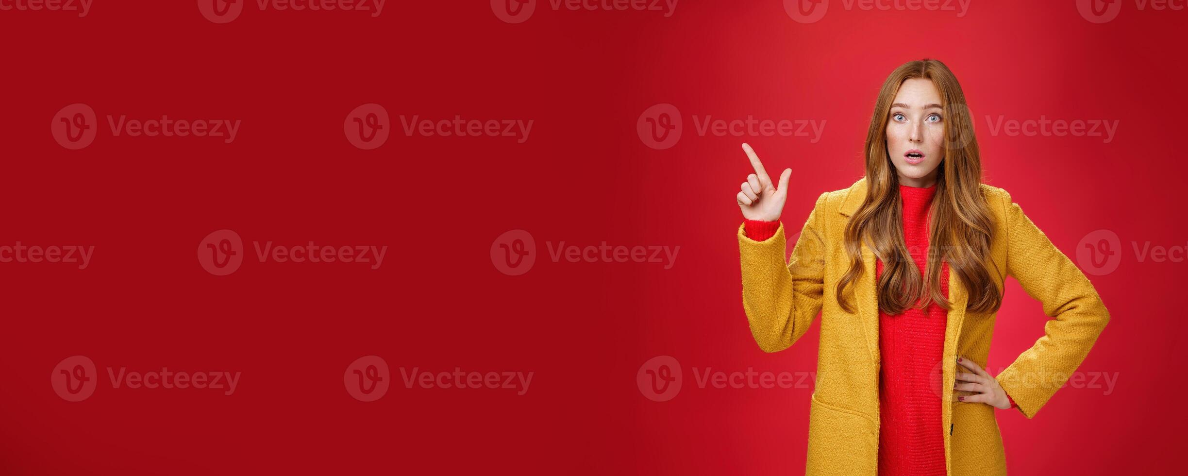 Shocked and stunned cute, confused ginger girl in yellow coat feeling worried and surprised open mouth lifting eyebrows amazed pointing at upper left corner, posing against red background photo