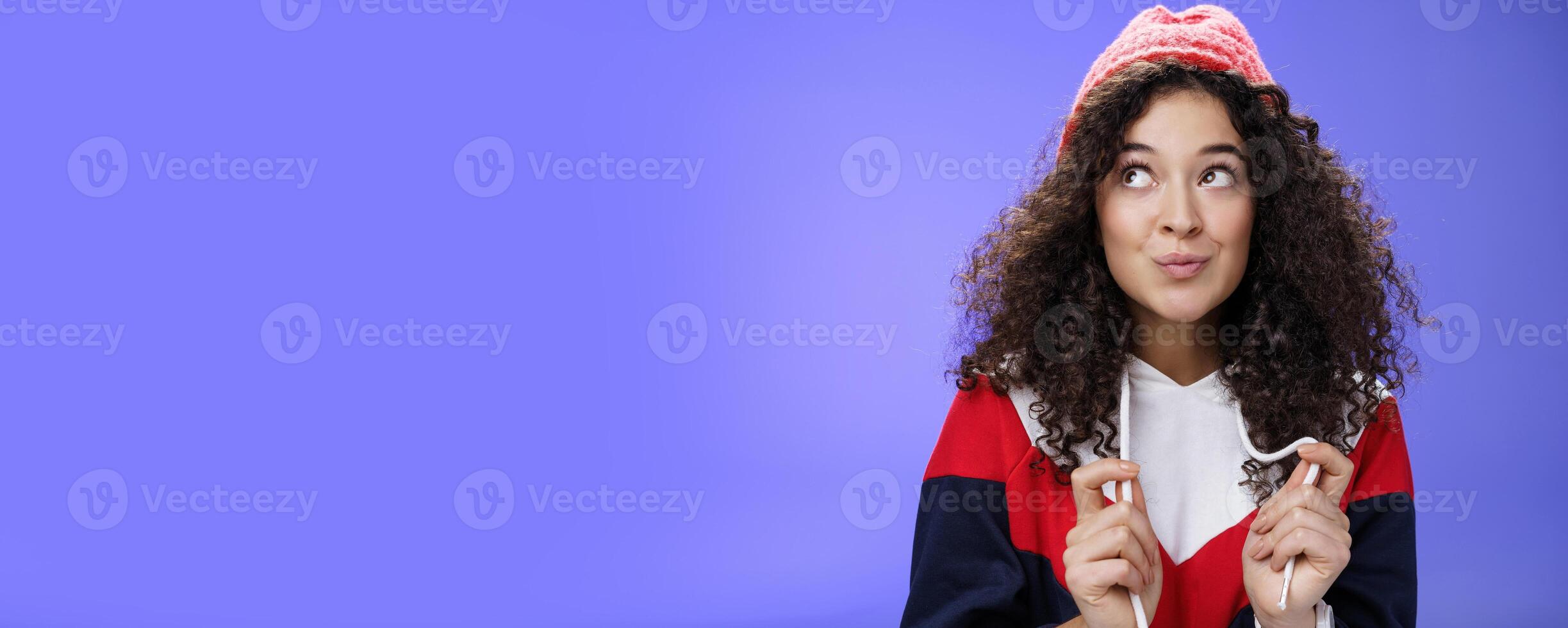 Sweet and silly cute 20s girl with curly hairstyle in warm red hat folding lips flirty and gazing at upper left corner sensually and dreamy posing against blue background playfully photo