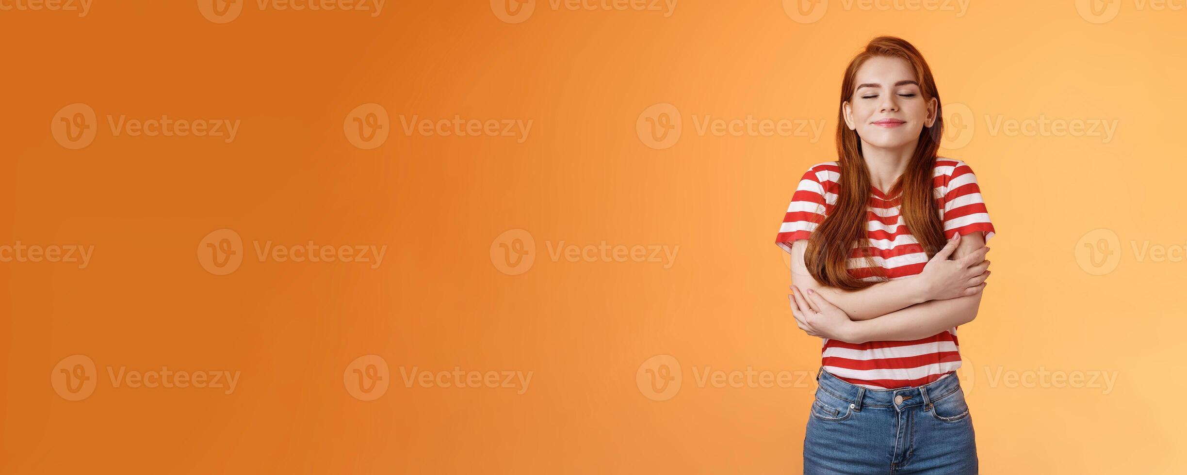 Tender lovely redhead modern woman embrace own body, feel happiness self-acceptance, smiling close eyes, cross arms, hugging herself dreamy, imaging something, daydreaming orange background photo