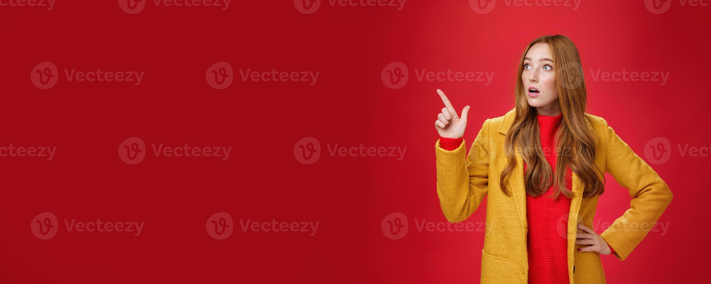 Portrait of empathic shocked and surprised stunned redhead woman in yellow coat open mouth amazed and speechless pointing and looking at upper left corner concerned and shook over red wall photo