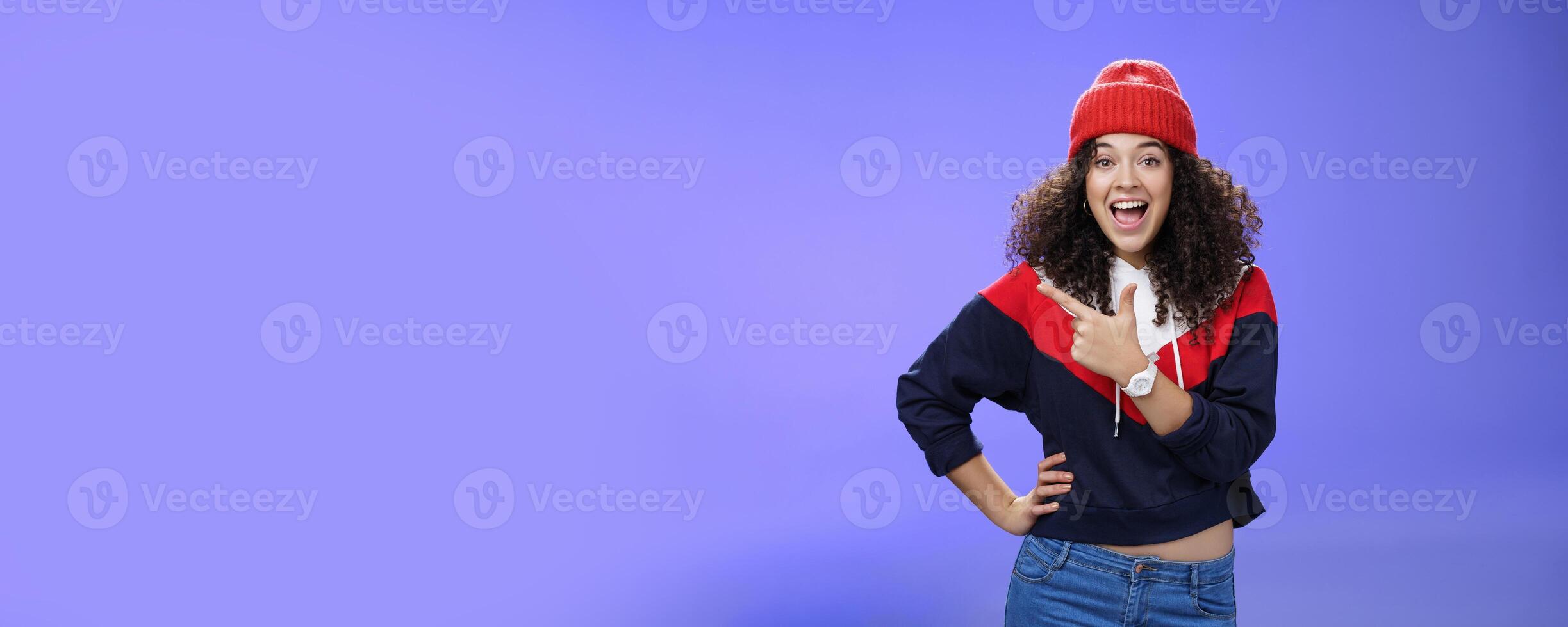 Portrait of enthusiastic and sociable european woman in warm hat and sweatshirt smiling delighted with amused grin as pointing at upper left corner impressed and astonished with awesome promotion photo