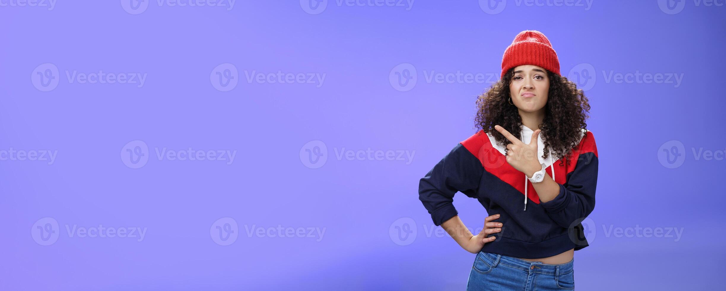 Girl feleing sad missing good chance, such shame. Portrait of displeased woman regretting frowning and pursing lips gloomy pointing at upper left corner upset wearing warm beanie over blue wall photo