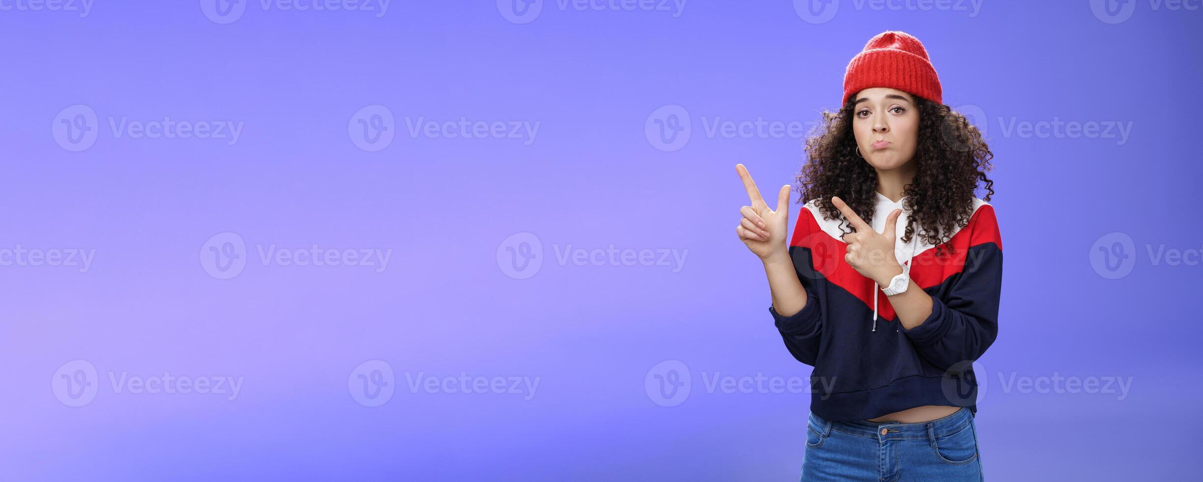 Portrait of silly upset cute curly-haired woman in trendy beanie pursing lips in sadness and frowning making disappointed, regret face pointing at upper left corner unhappy and sad over blue wall photo