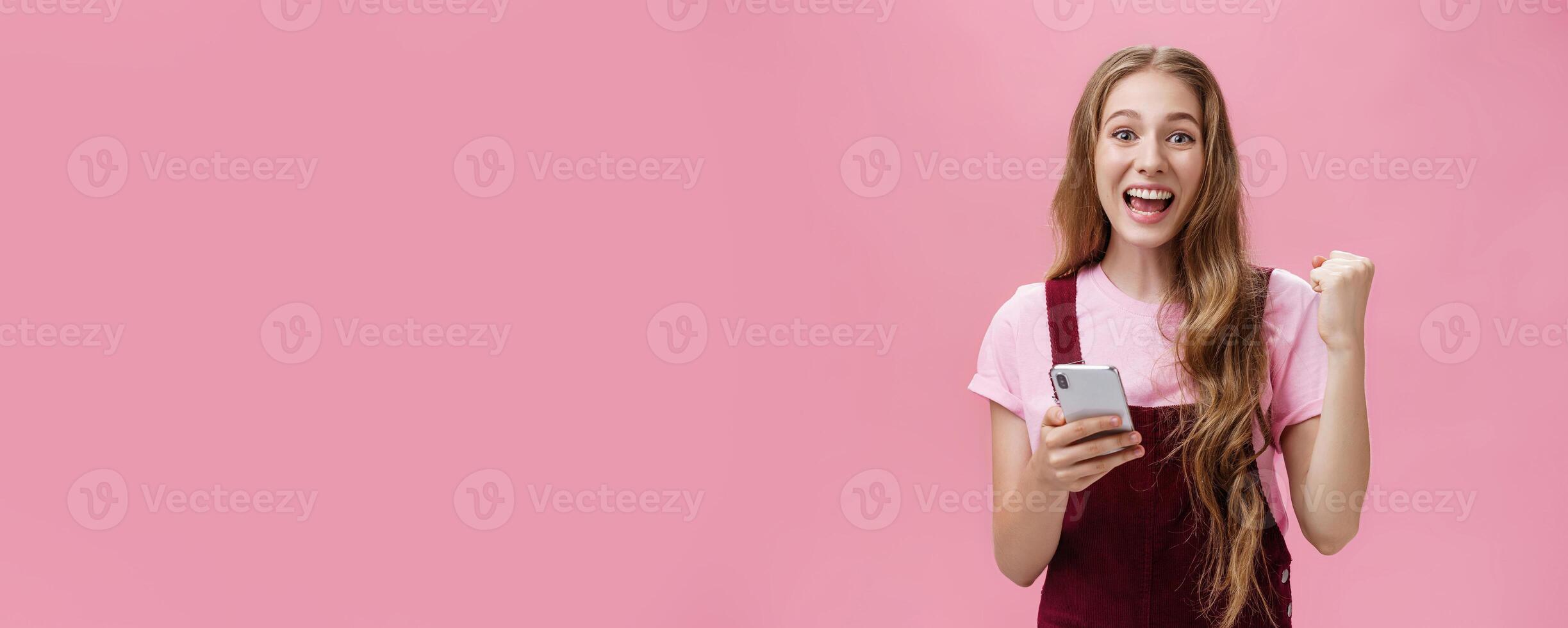 Yes I did it. Happy young excited cute woman in trendy outfit expressing cheer and joy smiling happily at camera raising fist in triumph and success holding smartphone beating own record in game photo