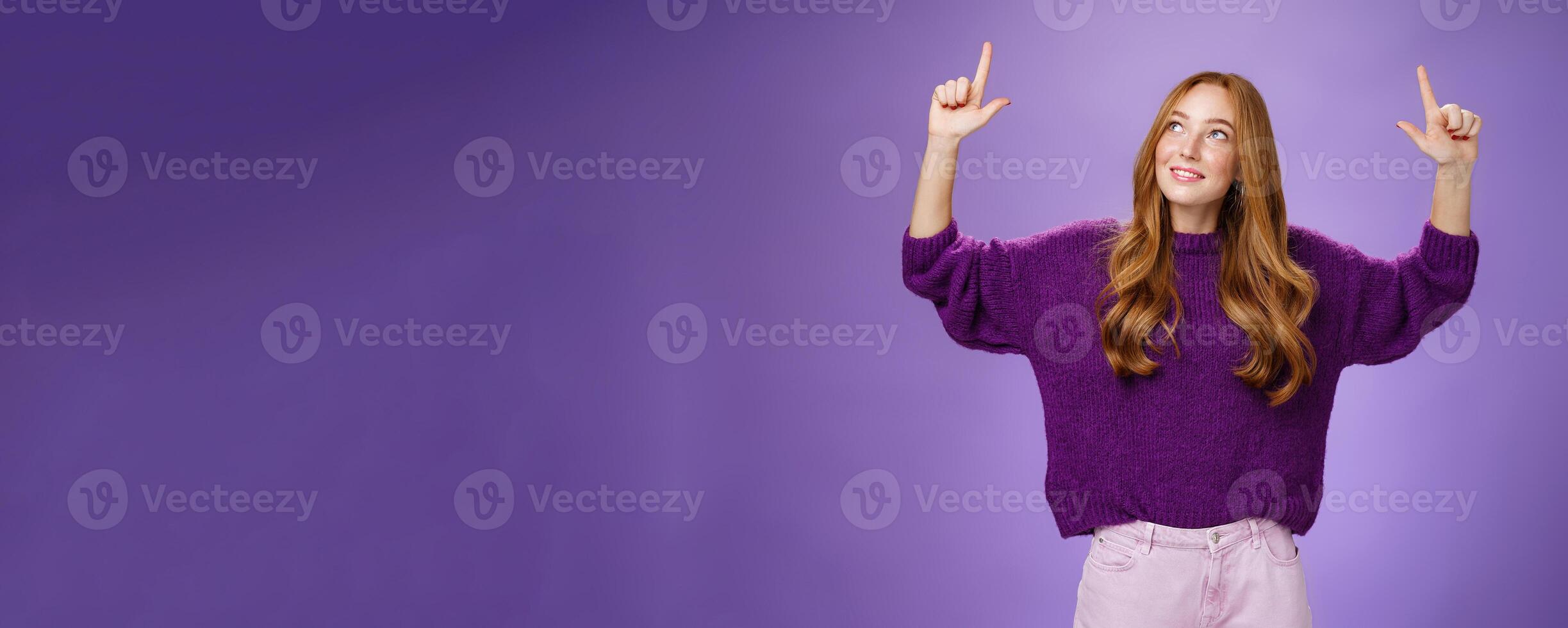 Dreamy girl with red hair and freckles in warm cozy purple sweater raising hands looking and pointing up with intrigued and happy charmed expression smiling as liking curious product over violet wall photo