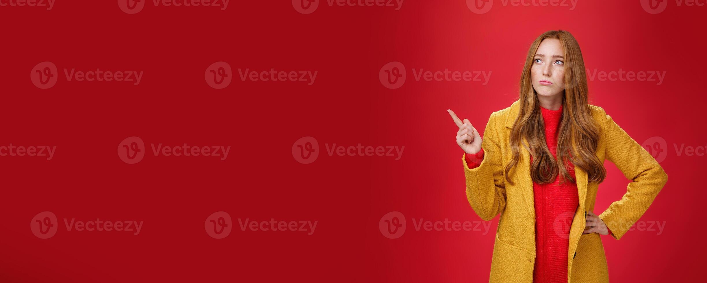 Sad and gloomy cute ginger girl in 20s wearing trendy yellow coat holding hand on waist, pouting, looking and pointing at upper left corner with regret and sorrow over red background photo