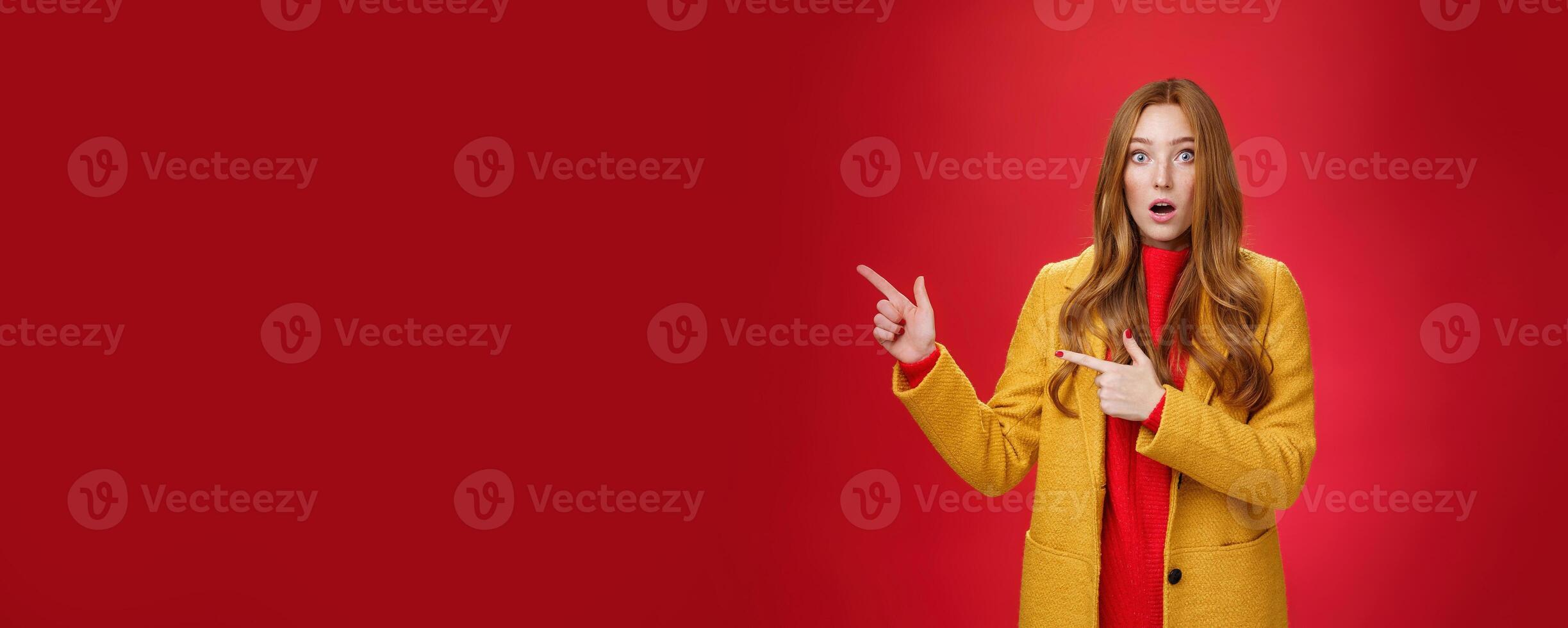 Stunned speechless and impressed attractive redhead woman with freckles in yellow coat drop jaw amazed as popping eyes at camera questioned, pointing left astonished over red background photo