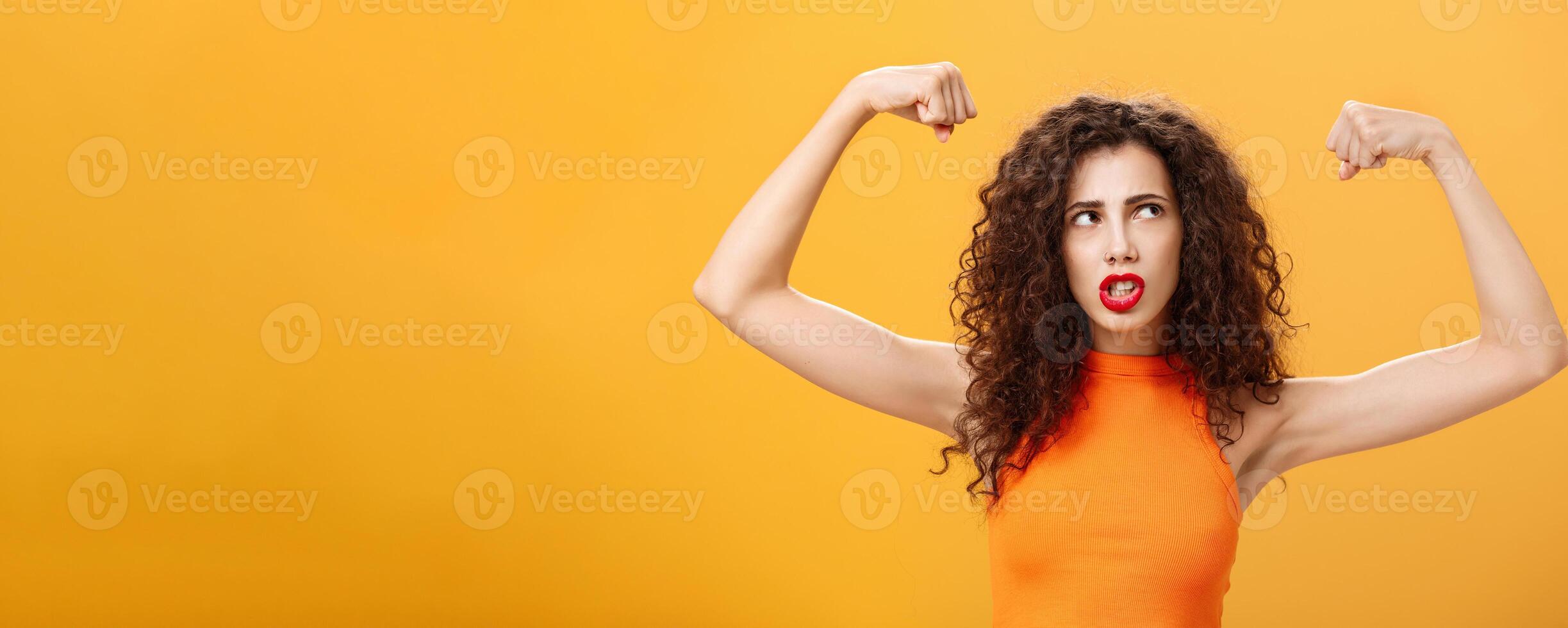Woman feeling powerful and strong raising hands with clenched fists making intense face being working out in gym showing muscles and biceps looking at upper right corner posing over orange background photo