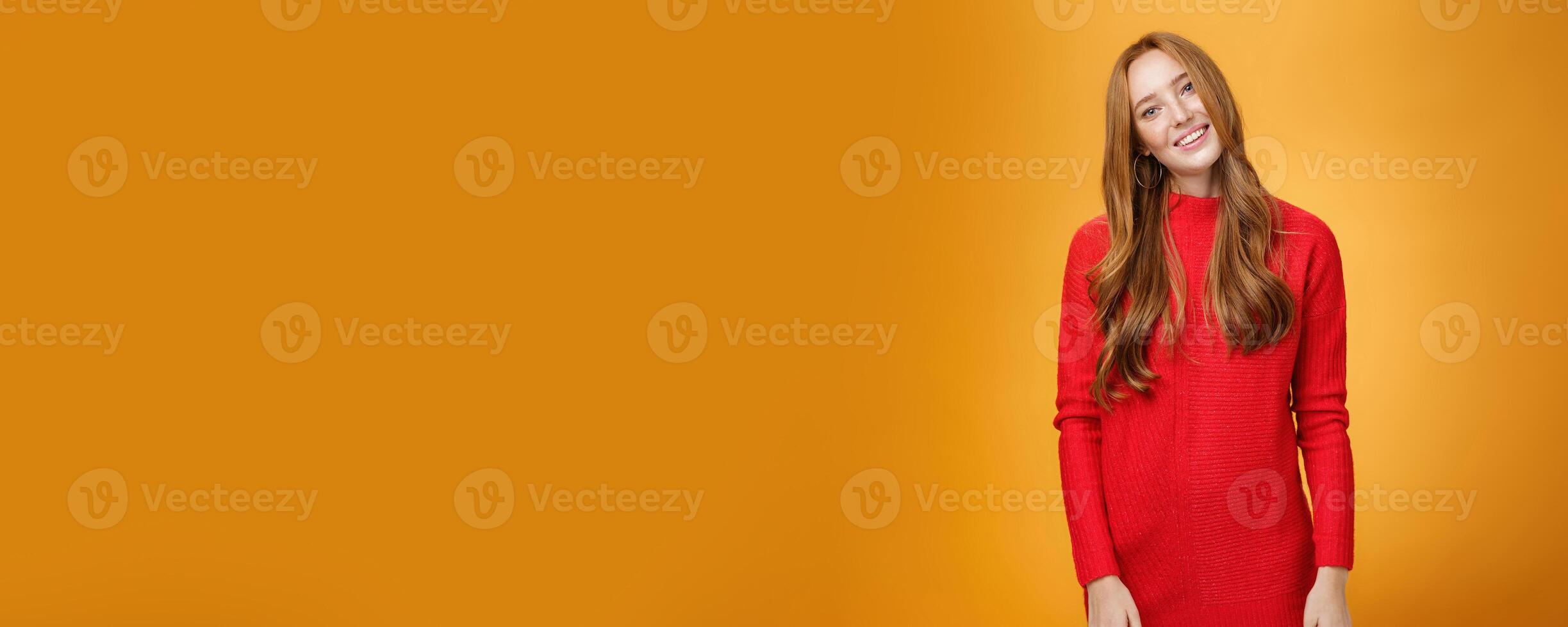 Attrative and stylish elegant busiensswoman with ginger hair in red knitted dress tilting head and smiling flirty and happily posing over orange background as listening with interest to conversation photo