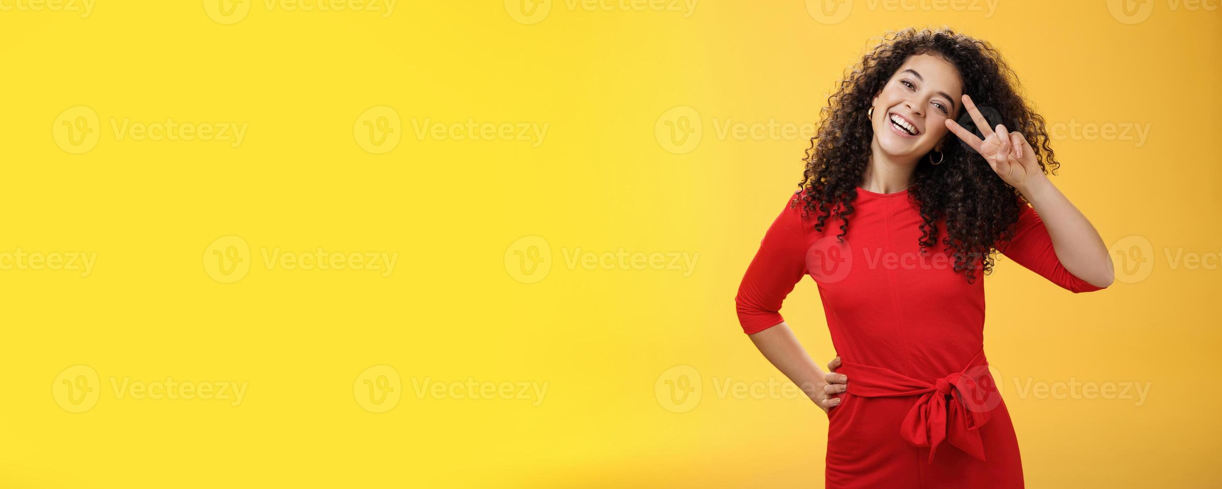 Waist-up shot of friendly tender and feminine cute woman with curly hairstyle tilting head and smiling, laughing and showing victory or peace gesture at camera over yellow background photo