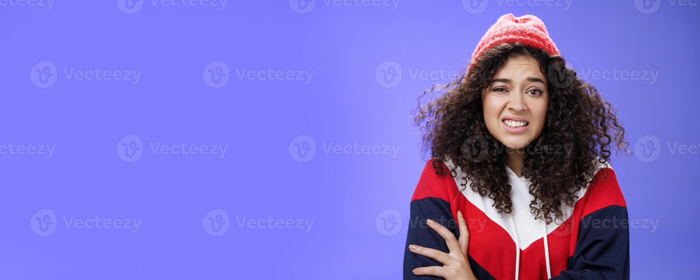 Woman with curly hair in winter beanie feeling uncomfortable and discomfort clenching teeth and frowning intense as hugging herself insecure and awkward, unwilling to say cruel rejection, feel awkward photo