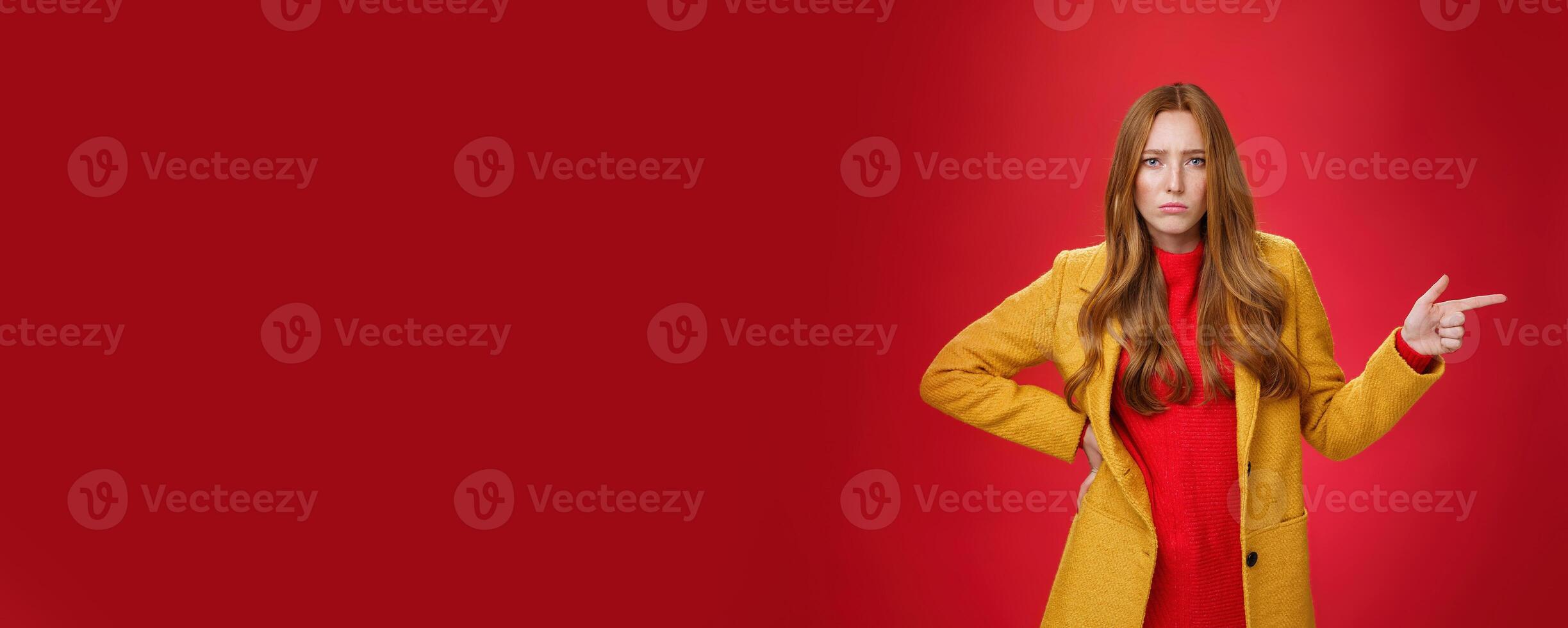 Explain yourself now. Portrait of upset gloomy and sad cute redhead girl waiting explanations asking question, pouting offended and pointing right with finger looking unhappy at camera over red wall photo