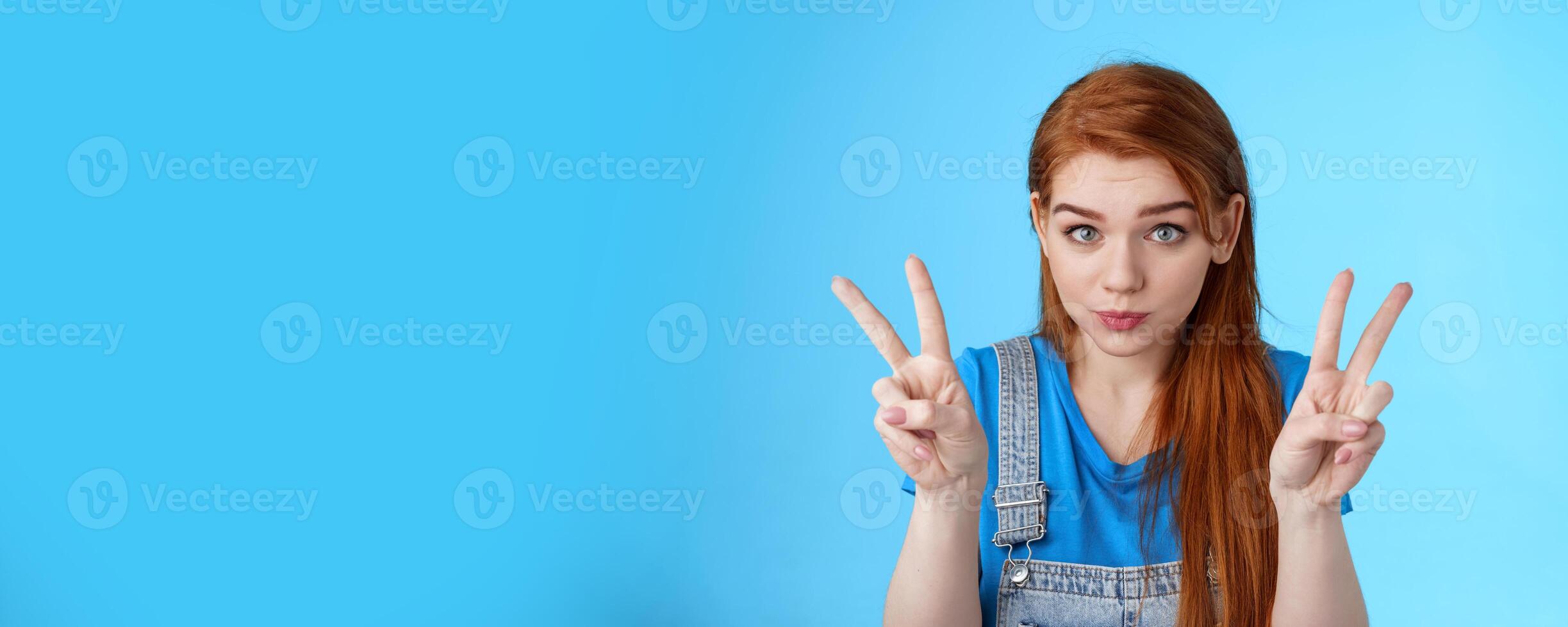 Playful cute silly redhead girlfriend making funny face, show peace victory signs, hold breath, pouting childish, fool around having fun, stand blue background, relaxing entertain siblings photo