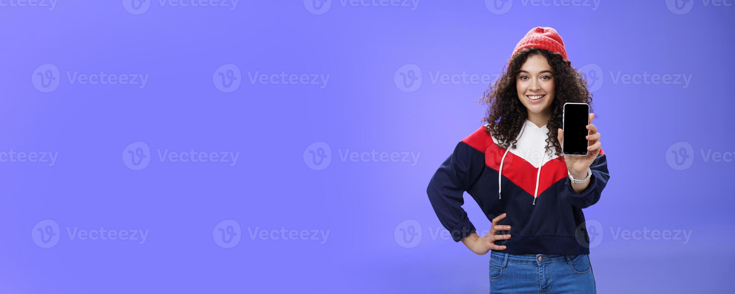 Girl showing her post as pulling smartphone screen towards camera smiling delighted waiting opinion, holding hand on waist in confident pose wearing warm winter red beanie and sweatshirt photo
