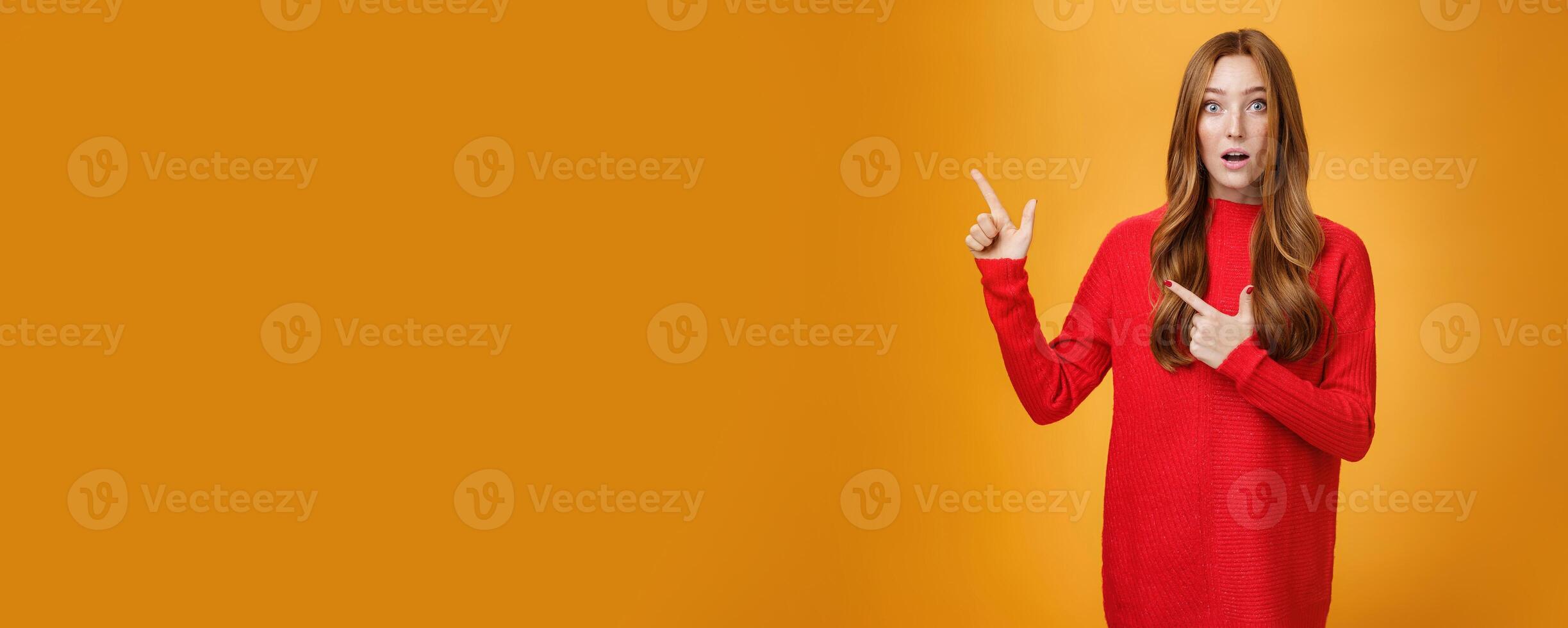 Surprised and impressed intrigued nice cute redhead female with freckles in warm red sweater pointing at upper left corner open mouth from amazement and looking questioned at camera over orange wall photo