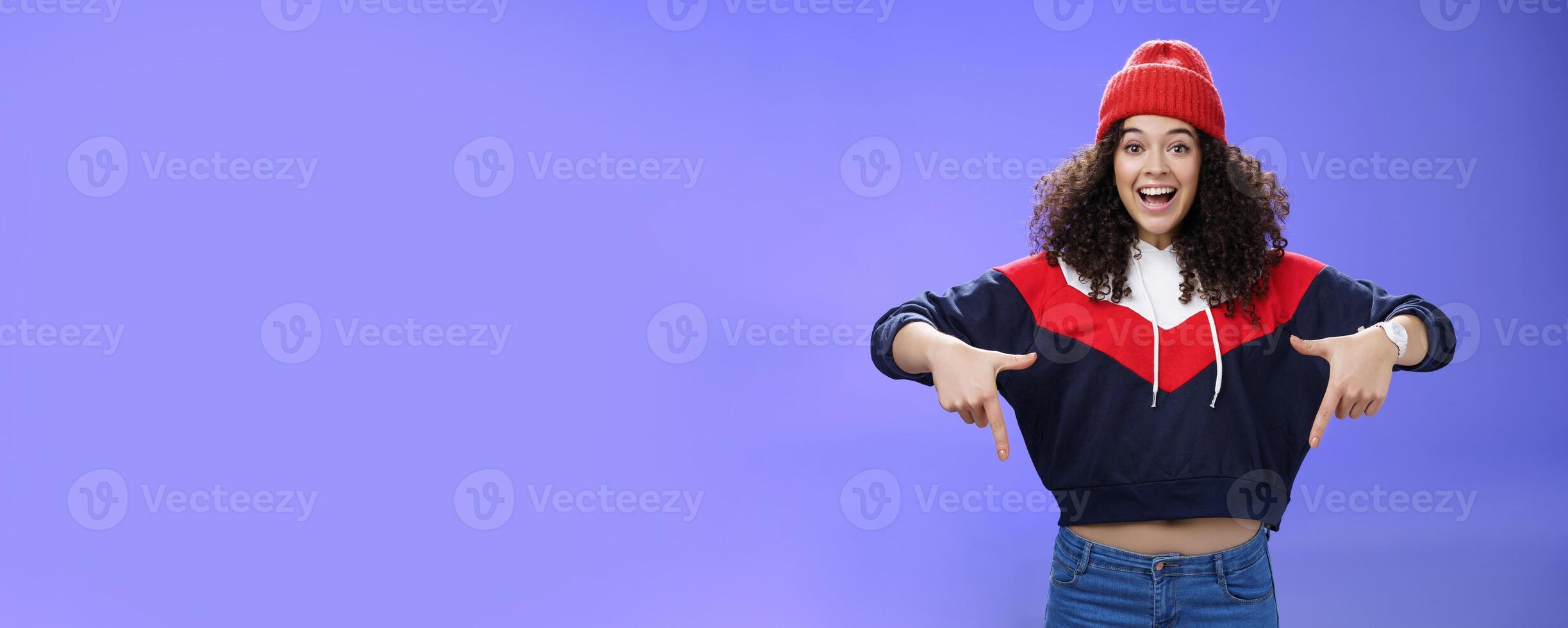 Hurry up and chek it out. Portrait of charismatic joyful and excited young cute curly-haired girl in beanie and stylish outfit pointing down, smiling broadly inviting join fun over blue background photo