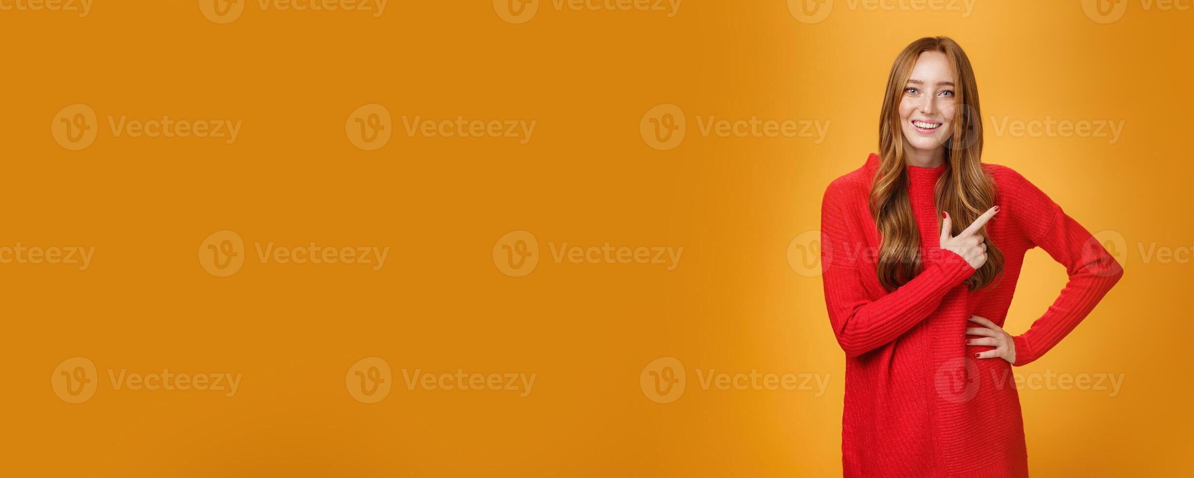 Attractive redhead successful woman in knitted warm dress pointing at upper right corner smiling cute and joyful at camera as asking question, having conversation over orange wall photo