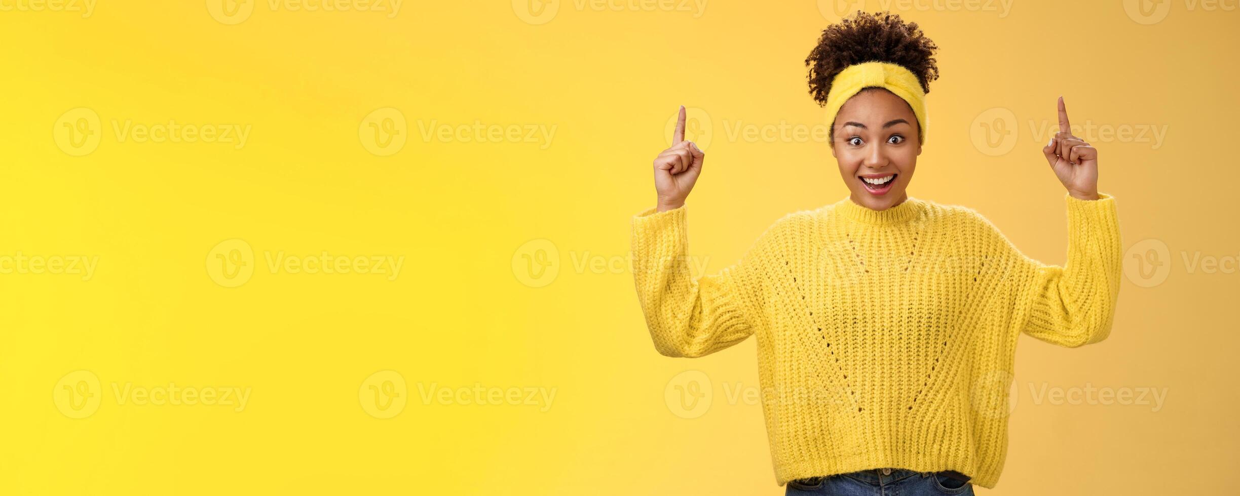 Charming emotive young african-american woman in sweater headband winning impressive sum money pointing up index fingers showing link astonished surprised smiling broadly yellow background photo
