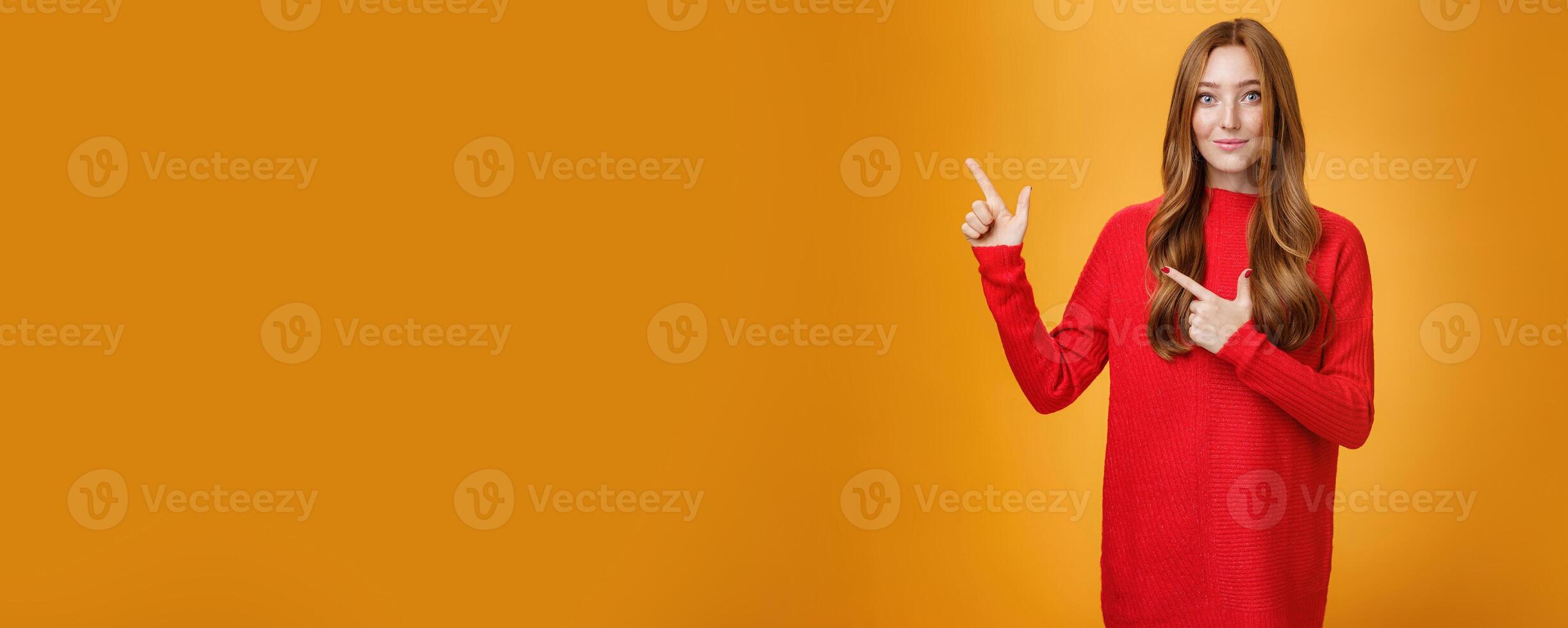 Soft and gentle gorgeous ginger girl with freckles in red warm sweater smiling joyfully pointing at upper left corner smiling showing curious place, suggesting visit it over orange background photo