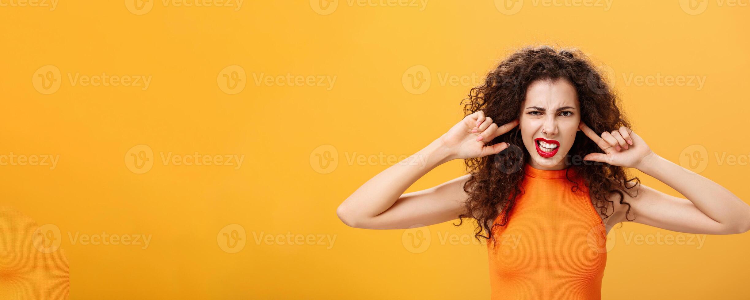 Waist-up shot of irritated bothered stylish woman with curly hair and red lipstick squinting clenching teeth waiting for loud bang covering ears with index fingers being annoyed of noisy neighbours photo