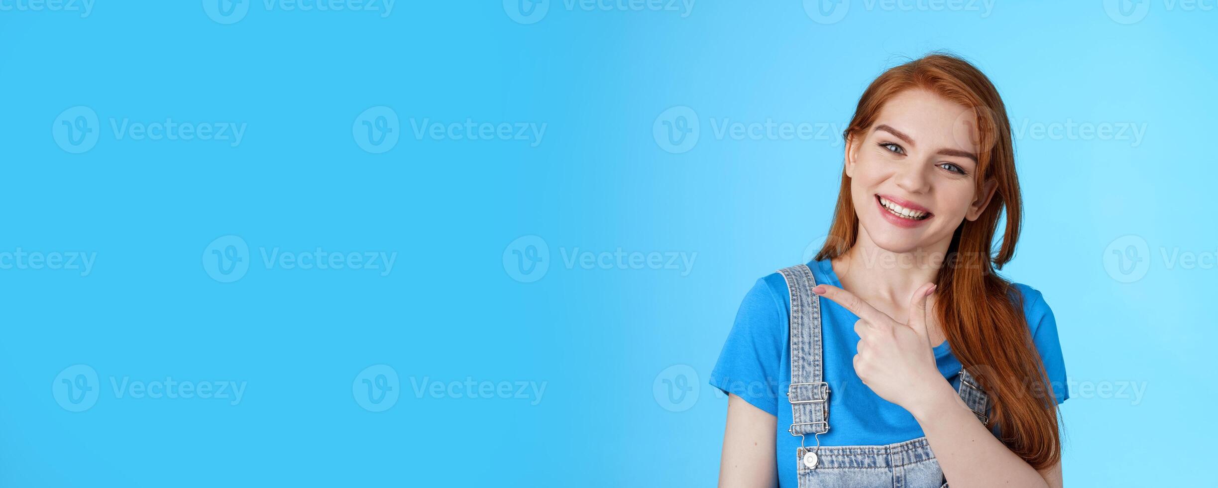 Friendly cheerful caucasian red woman, tilt head smiling pleasantly, suggest good copy space, pointing left introduce promo, give advice, indicating awesome link, pointing blue background photo