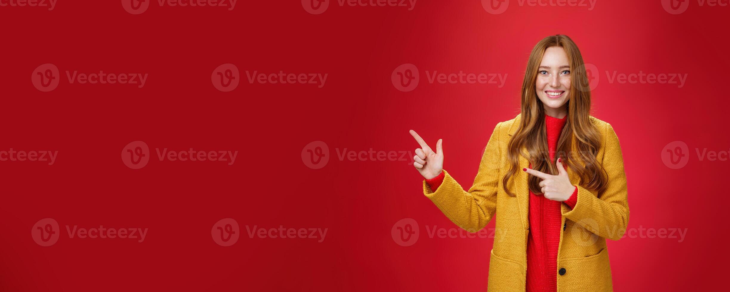 Cheerful friendly-looking and energized helpful redhead girl in yellow coat showing way pointing at upper left corner and smiling broadly with satisfied grin posing delighted over red background photo