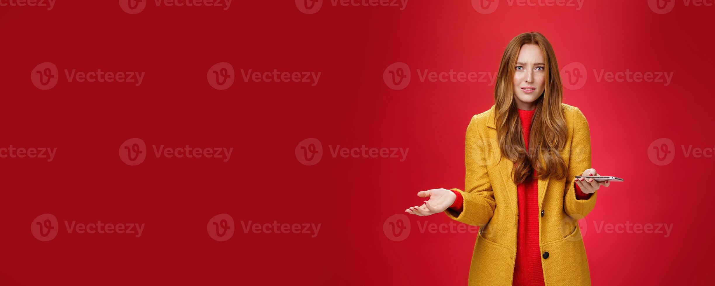 What a heck going on. Portrait of annoyed and confused perplexed ginger businesswoman in yellow coat shrugging with raised palm in clueless gesture holding smartphone troubled and questioned photo