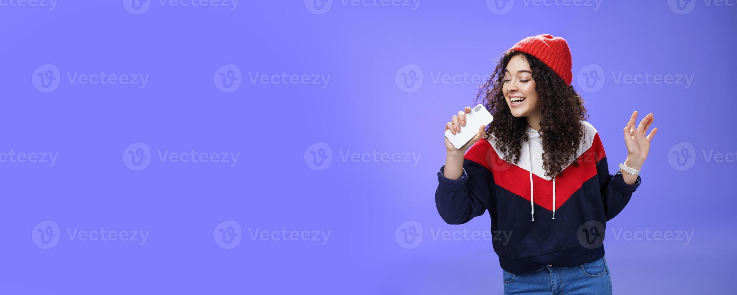 Attractive happy young woman with curly hair in hat singing enjoying perfect winter day singing along in smartphone, holding mobile phone like microphone, adore karaoke over blue background photo