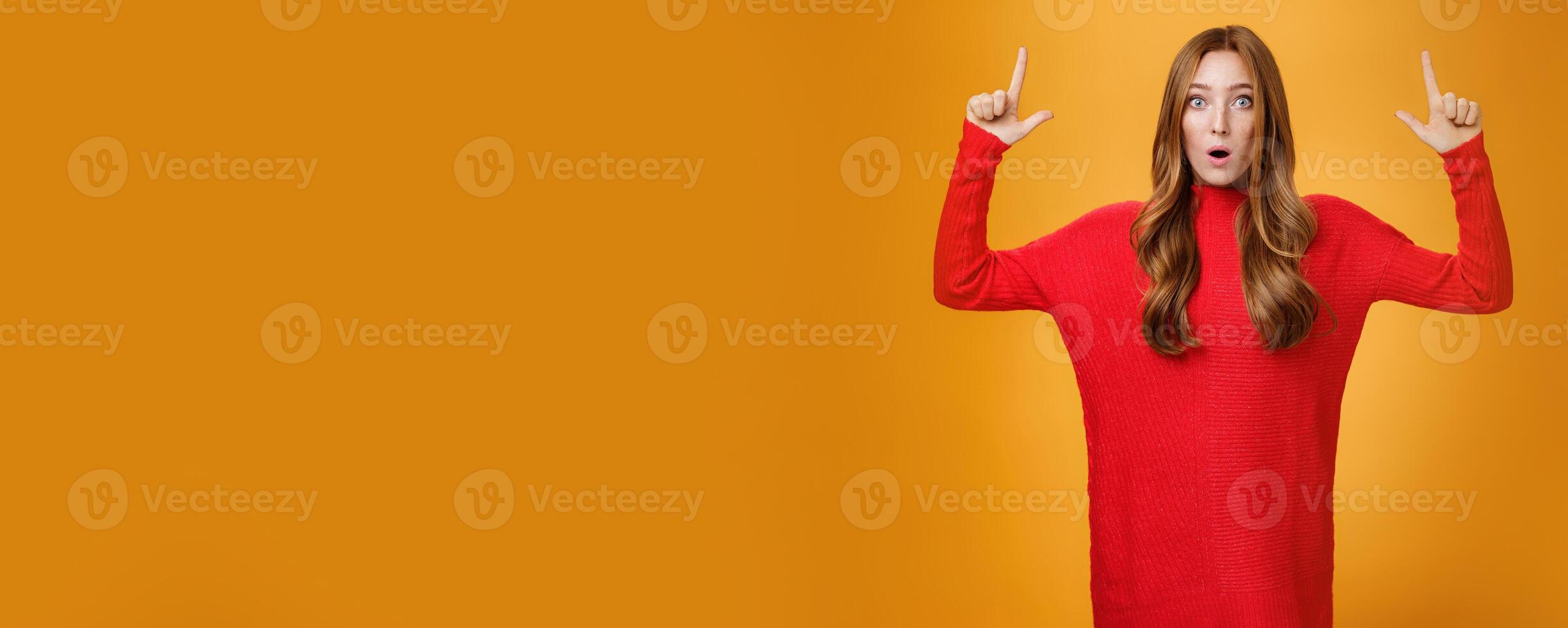 Portrait of attractive and stylish redhead 25s female in knitted red dress gasping from amazement open mouth questioned and surprised as pointing up at astonishing promotion over orange wall photo
