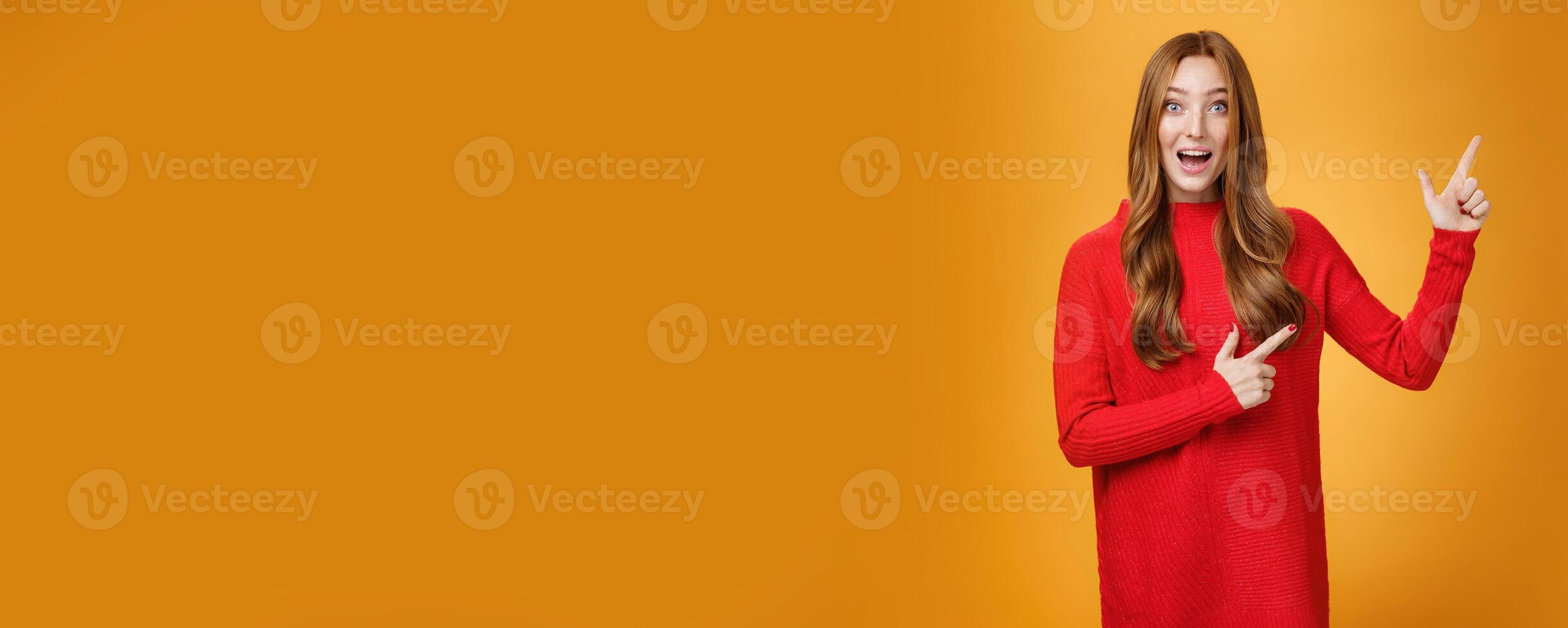 Enthusiastic and ambitious cute redhead female with freckles in red dress pointing at upper right corner smiling amazed and curious as exploring awesome new space posing over orange background photo