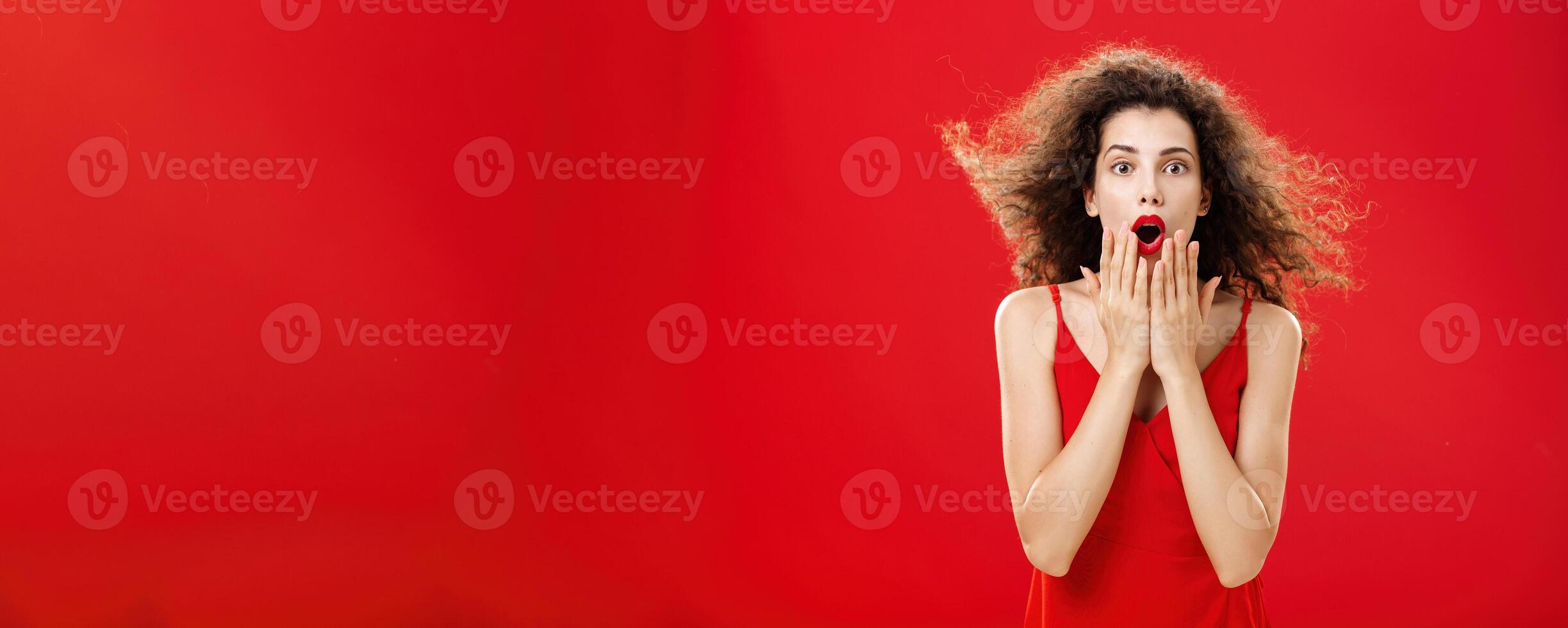 Attractive elegant young woman in red evening dress with curly hair flicking on wind saying wow being impressed and thrilled covering opened mouth from amazement standing over red background photo