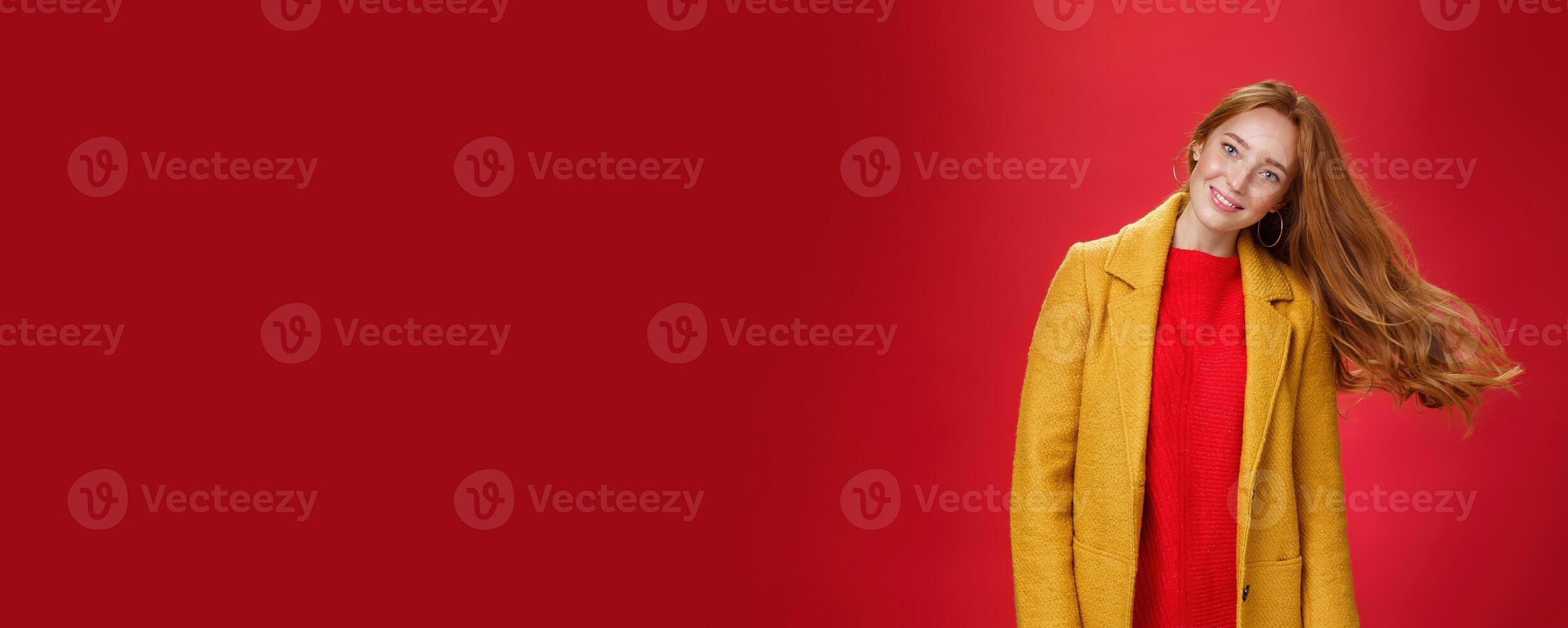 Charming redhead female with freckles waving hair smiling broadly tilting head as haircut flying in air posing delighted and carefree over red background in warm yellow coat and knitted dress photo