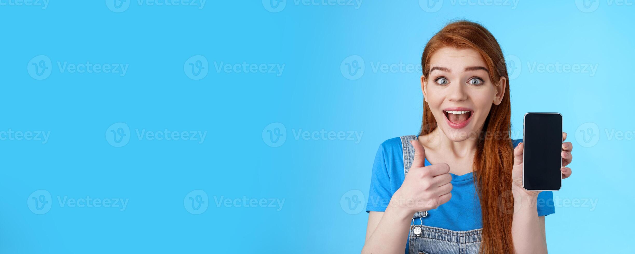 Amazed fascinated cute redhead woman recommend good smartphone, awesome new app, phone game, show cellphone display, look thrilled camera smiling approval, like cool device, blue background photo