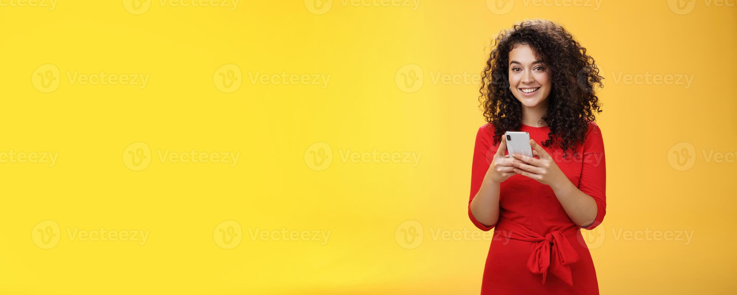 Girl messaging friend to tell all hot rumors holding mobile phone in hands smiling broadly and excited at camera as using smartphone browsing in net or edditing in app over yellow background photo