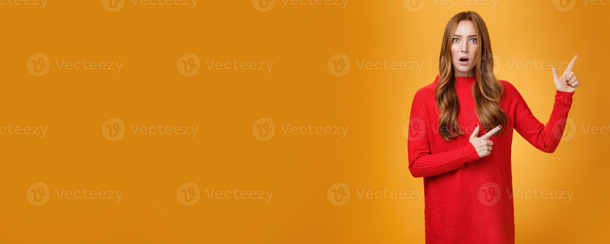Shocked distressed and confused ginger girl with freckles and blue eyes in red sweater pointing at upper right corner with shook and surprise open mouth, raise eyebrows from stupor over orange wall photo