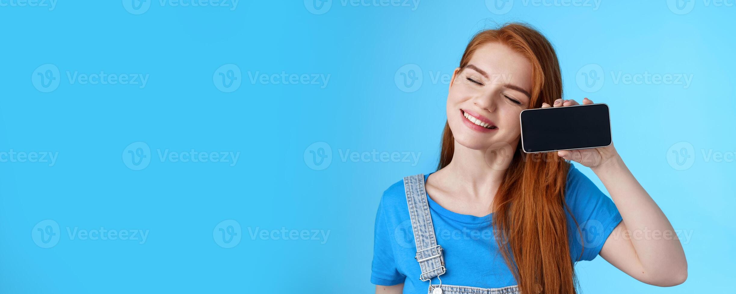 Carefree tender redhead european female tilt head, smiling closed eyes, hold smartphone horizontally, show loving girlfriend photos, romantic online, stand blue background cheerful photo