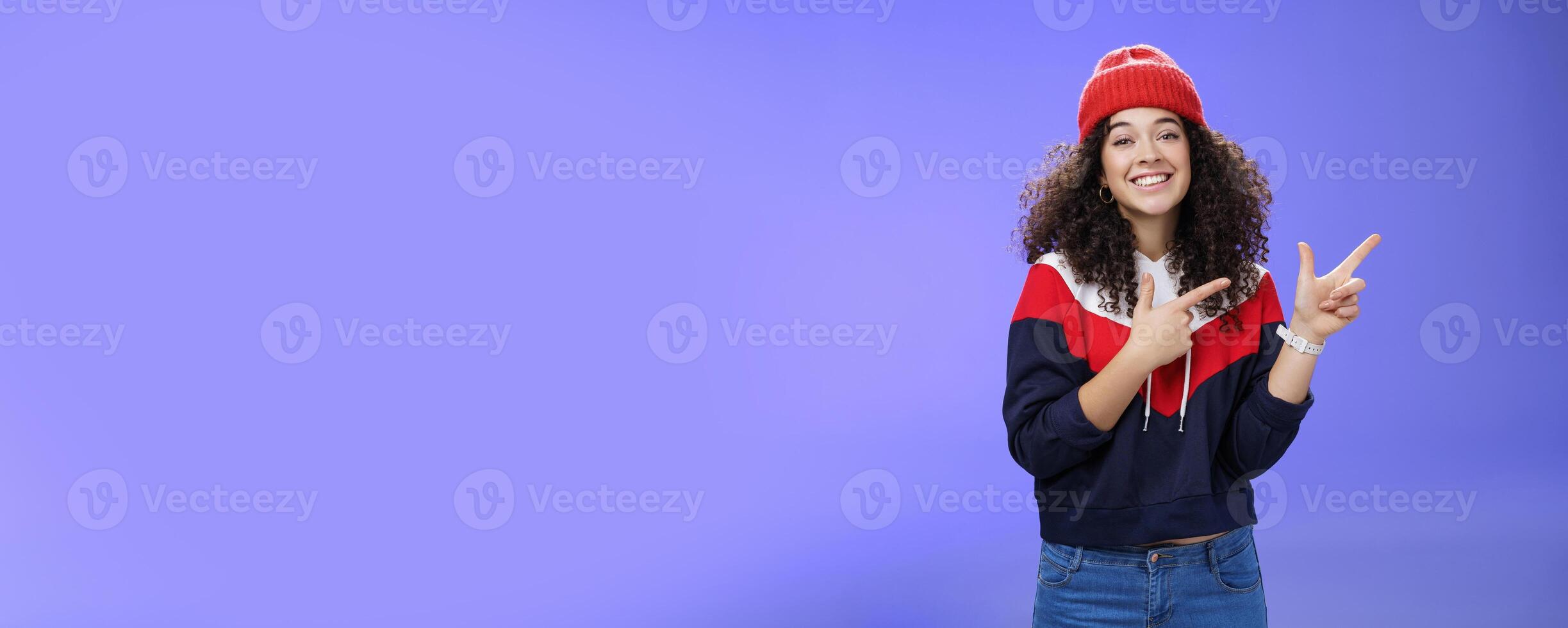 Cute female friend pointing at upper right corner with finger pistols as smiling broadly at camera wearing warm winter hat and sweatshirt skiing over blue background joyfully photo