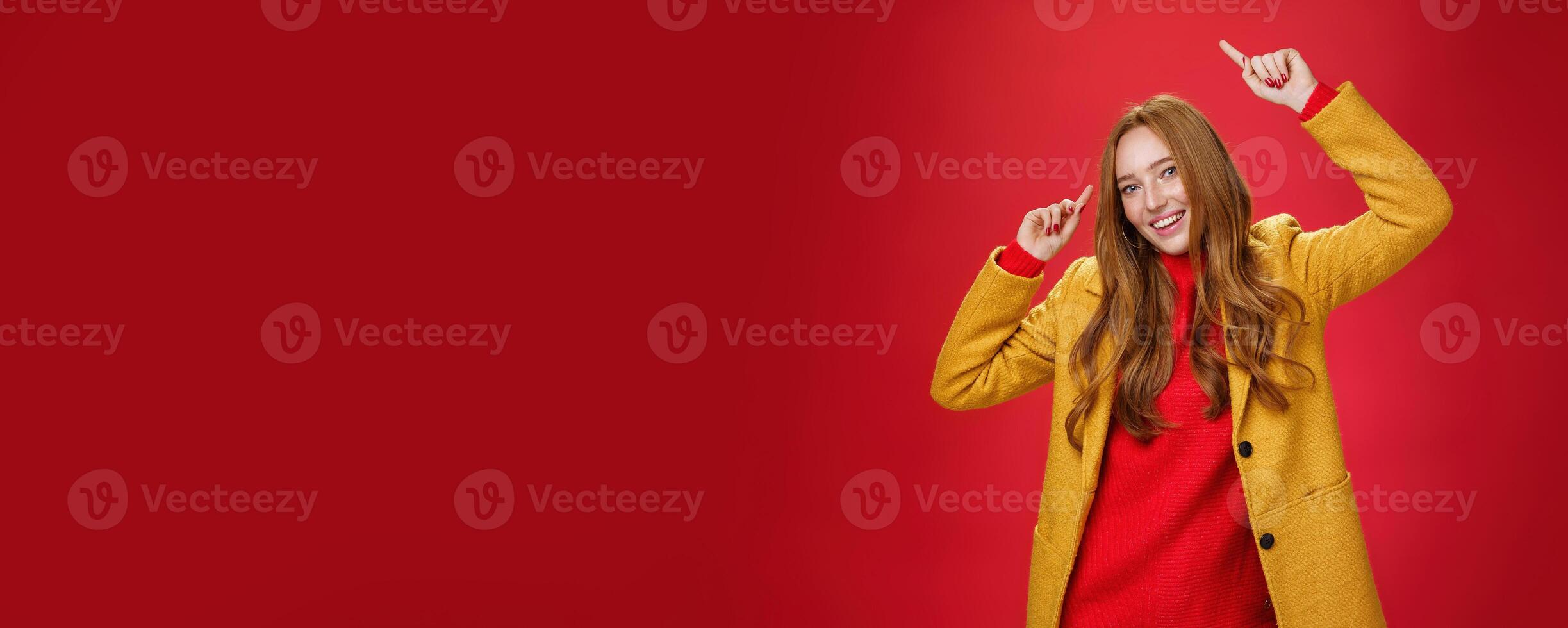 No time for sorrow dance and feel happy autumn came. Portrait of bright and cheerful charming redhead woman raising hands up moving to rhythm of music delighted posing in yellow coat over red wall photo