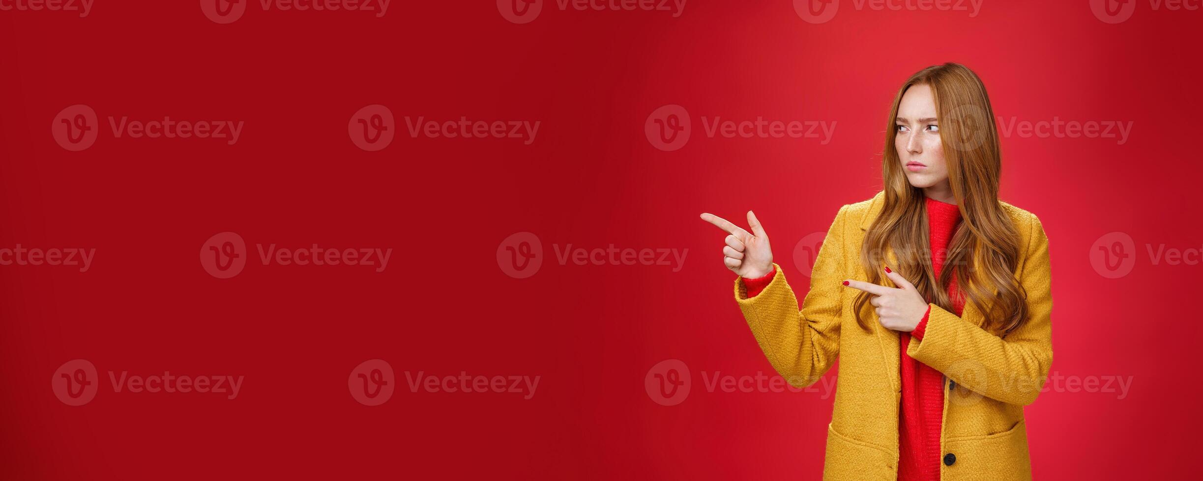 Studio shot of intense suspicious and displeased redhead female in yellow coat pointing and looking serious left squnting and frowning making focused look at something disturbing and doubtful photo