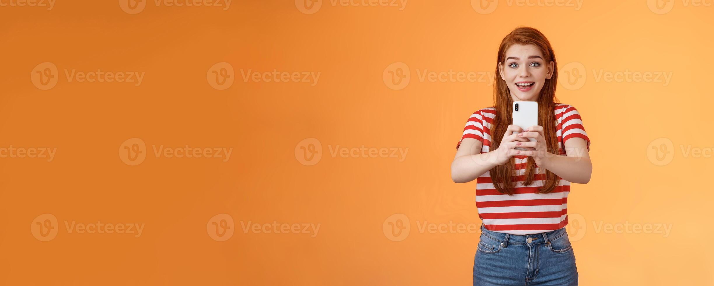 Amused excited redhead female record thrilled see famous person, smiling amazed hold smartphone taking picture, tourist adore trip shooting everything during summer journey, orange background photo