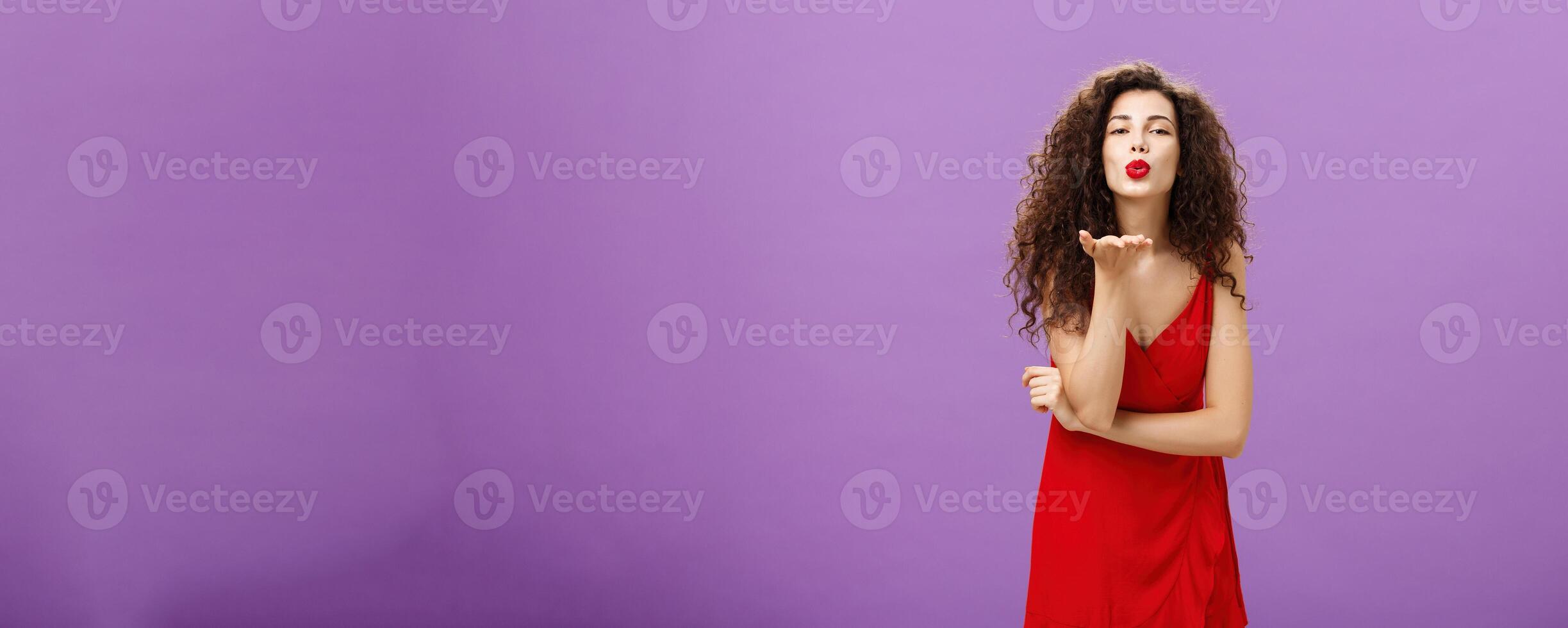 Woman sending passionate kiss being hopeless romantic standing in elegant red dress with curly hairstyle and makeup blowing mwah with smile and folded lips holding palm near mouth over purple wall photo
