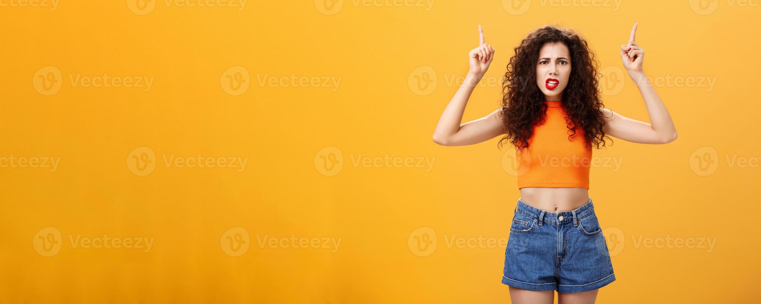 Indoor shot of displeased female client with curly stylish hairstyle red lipstick in cool cropped top pointing up demanding explanation with confused and dissatisfied expression over orange background photo