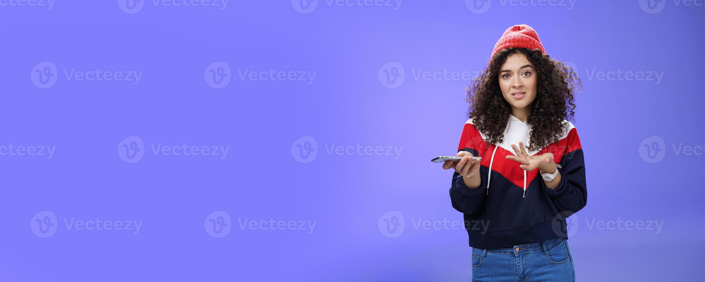 Confused and clueless girl cannot understand meaning of strange message shrugging grimacing questioned as holding smartphone and extending arm in unaware gesture over blue background photo