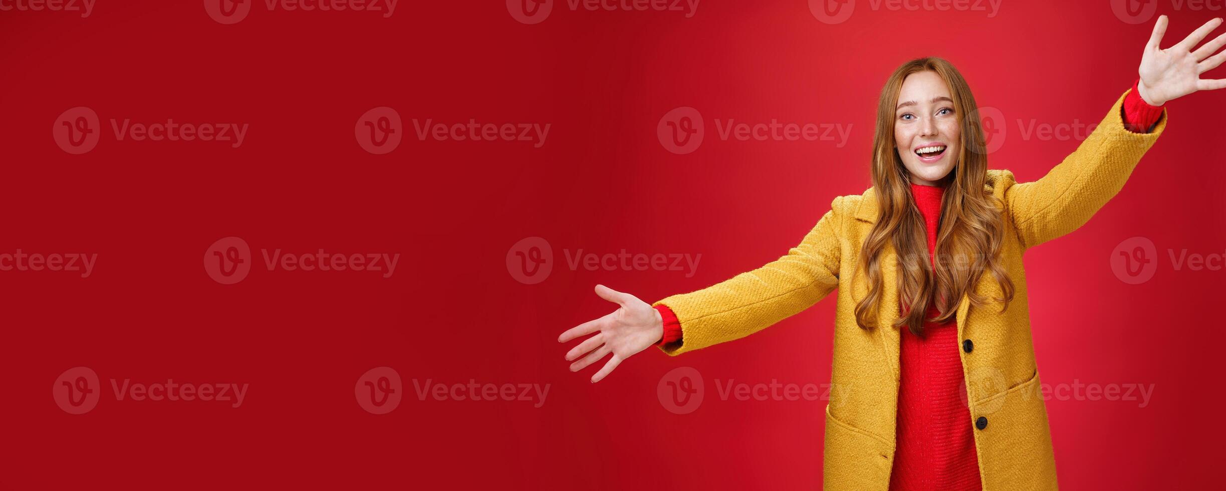 Hey come and hug me friend. Charming playful and emotional redhead female extanding palms forward to cuddle and greet, giving warm welcome smiling broadly posing in yellow coat over red wall photo