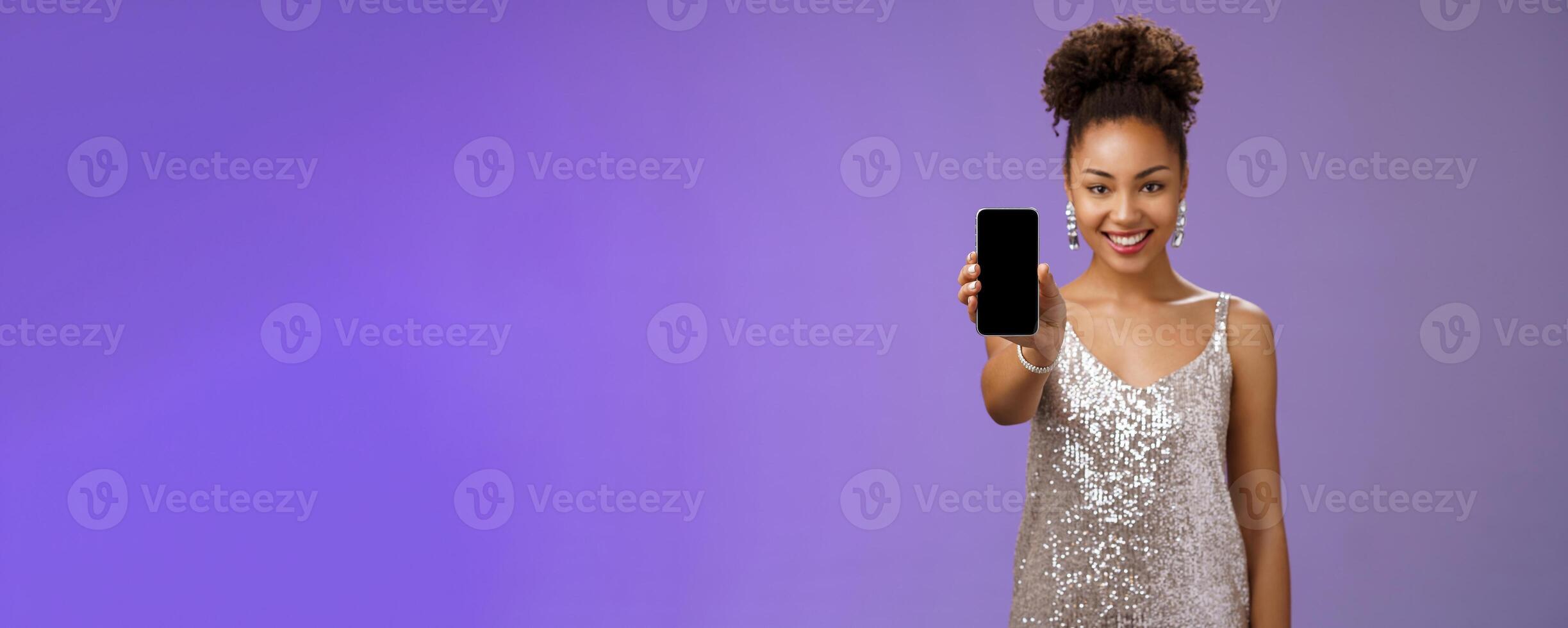 Charming attractive african-american woman in silver glittering stylish dress extend arm showing smartphone display presenting app promoting cool device standing pleased smiling blue background photo