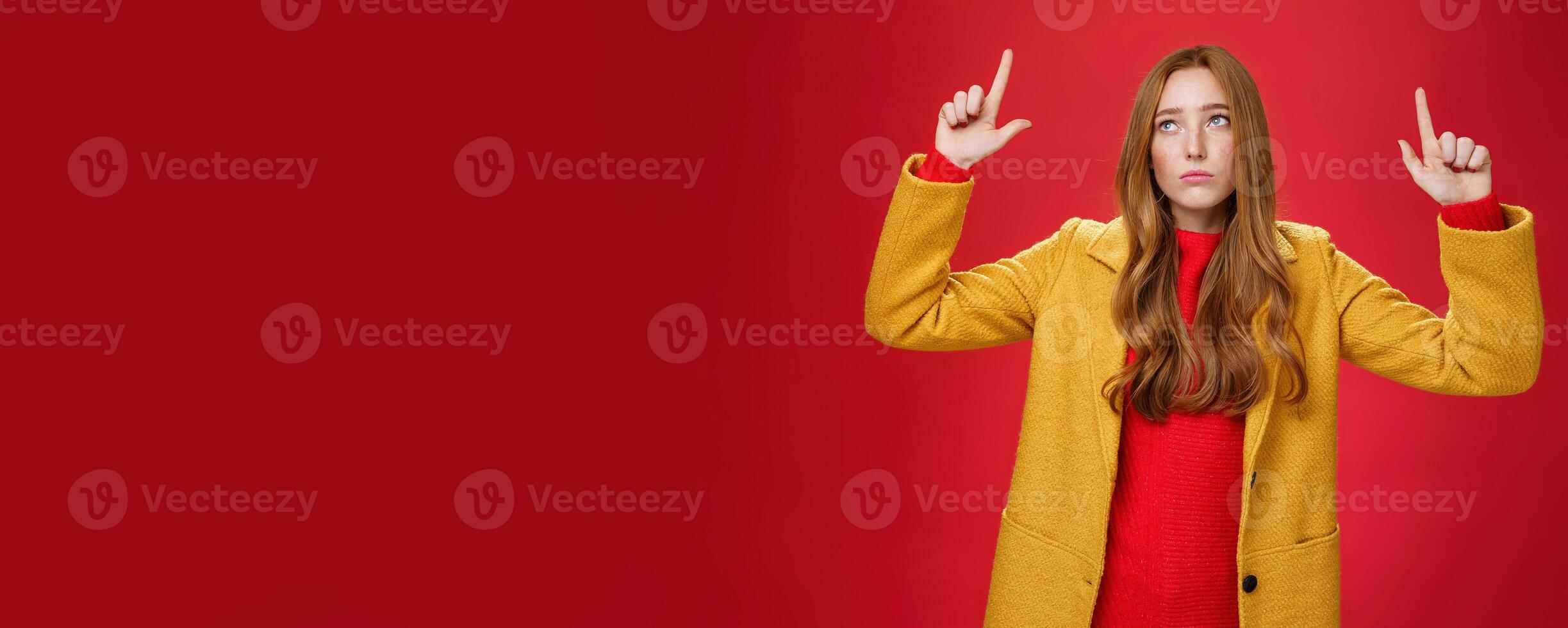 Sad and sorrow cute redhead female in yellow coat raising hands up displeased with regret looking at upper left corner apathical as being disappointed with rain or bad weather over red wall photo