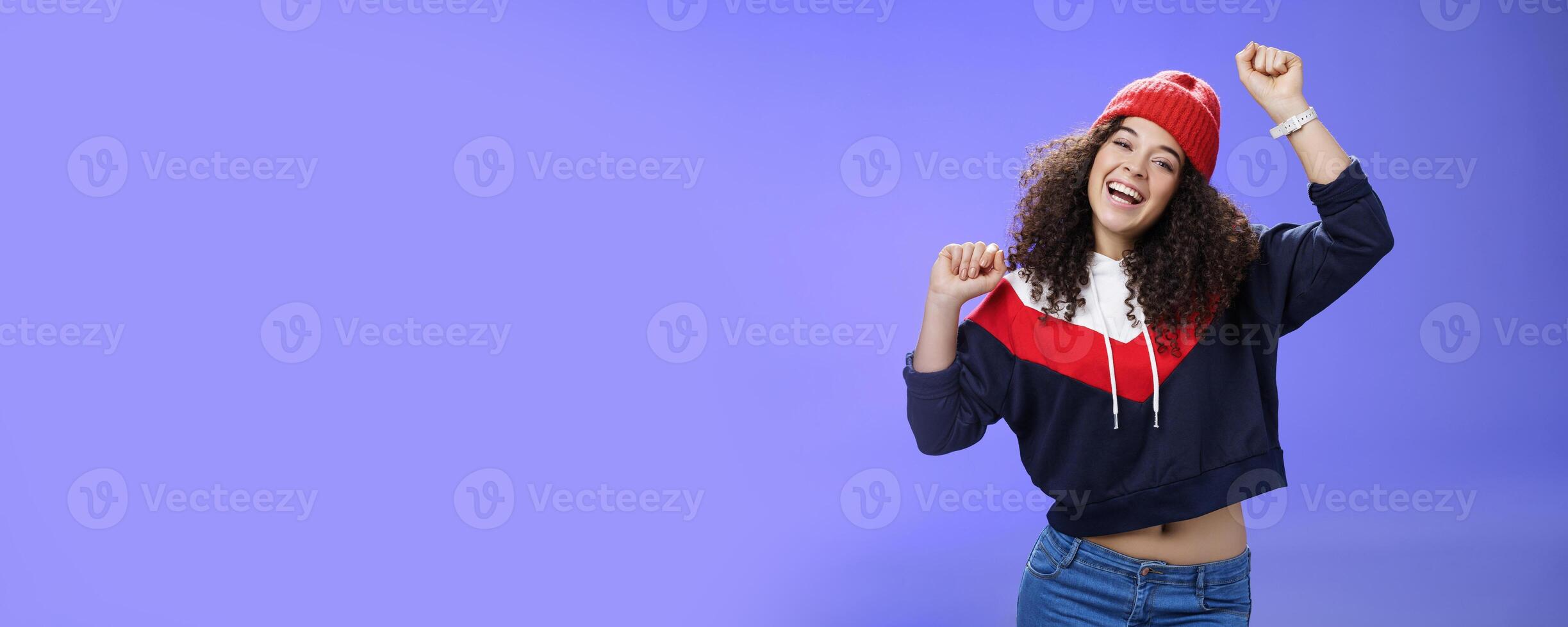 Hey come on dance with me. Friendly-looking bright and stylish cheerful woman with curly hairstyle wearing warm beanie raising hands up as enjoying great day, having fun outdoors over blue wall photo