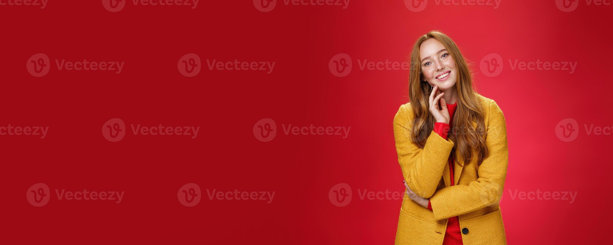 Good-looking carefree and happy relaxed redhead girlfriend in yellow stylish coat touching face and tilting head as smiling with positive emotions, posing cheerful against red background photo