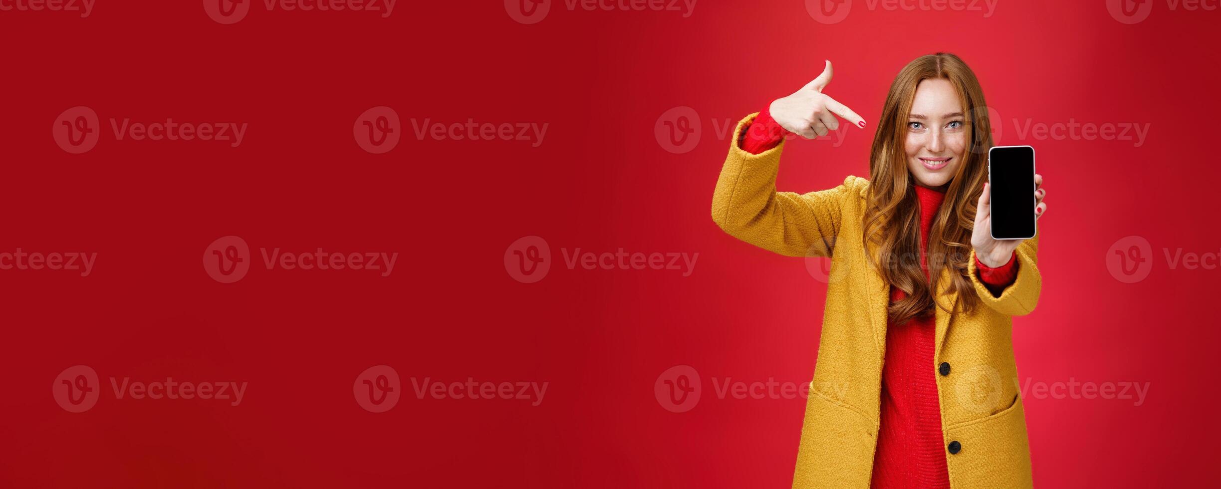 Pick this phone you never regret. Portrait of friendly-looking attractive and confident redhead glamour girlfriend in yellow coat showing smartphone pointing at mobile as smiling broadly at camera photo
